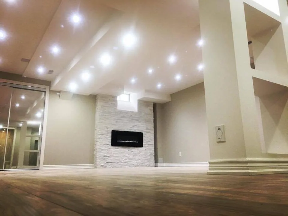 Finished basement with many recessed lights in a coffered ceiling, a stone feature wall with a wall-mounted electric fireplace and built-in shelving